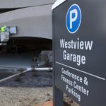 Two people in hardhats work in a crane on the side of the Westview Parking garage. In the foreground a sign says “Westview Garage Conference & Fitness Center Parking.”