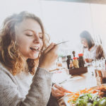 A woman sits at a table with other people. Food and condiments sit on the table. The woman bites food from a fork.