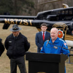 A man speaks at a podium in front of a helicopter