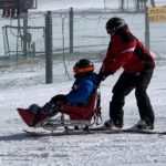 An instructor pushes an adaptive skier in a wheelchair down a slope at Roundtop Mountain Resort. Both are wearing ski jackets, ski pants and helmets. Two people stand in the background, and one person lies on the ground.
