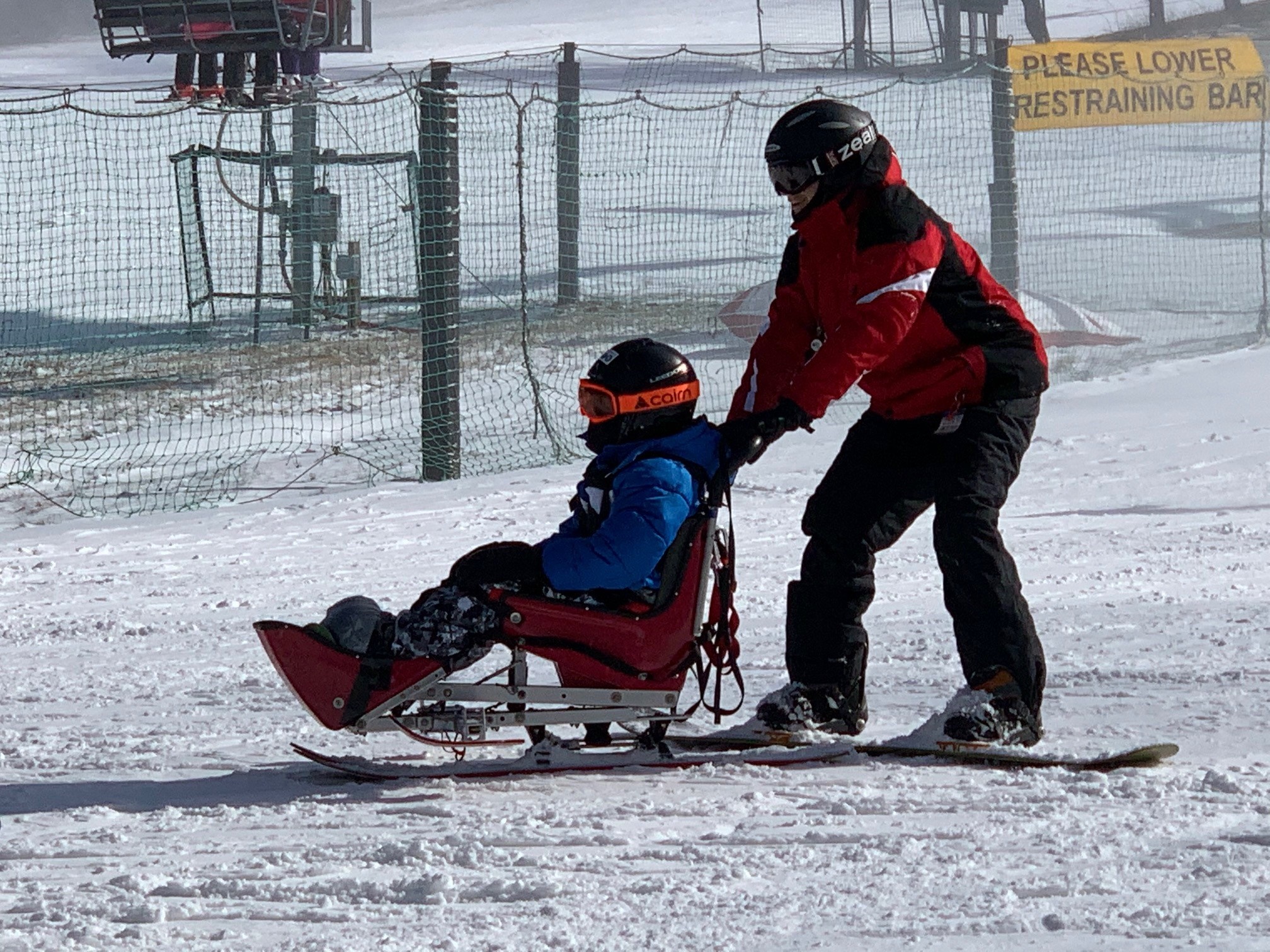 An instructor pushes an adaptive skier in a wheelchair down a slope at Roundtop Mountain Resort. Both are wearing ski jackets, ski pants and helmets. Two people stand in the background, and one person lies on the ground.
