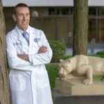 Kevin Black wears a lab coat and leans against a tree with his arms crossed. Behind him is a statue of the Penn State Nittany lion.