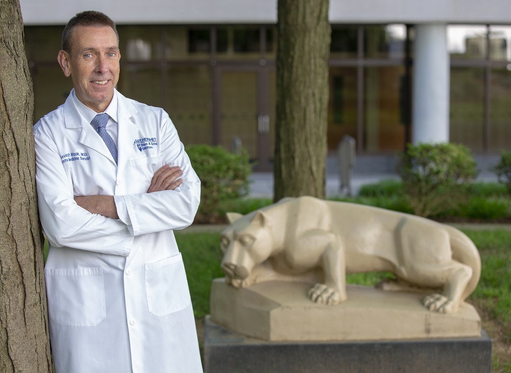 Kevin Black wears a lab coat and leans against a tree with his arms crossed. Behind him is a statue of the Penn State Nittany lion.