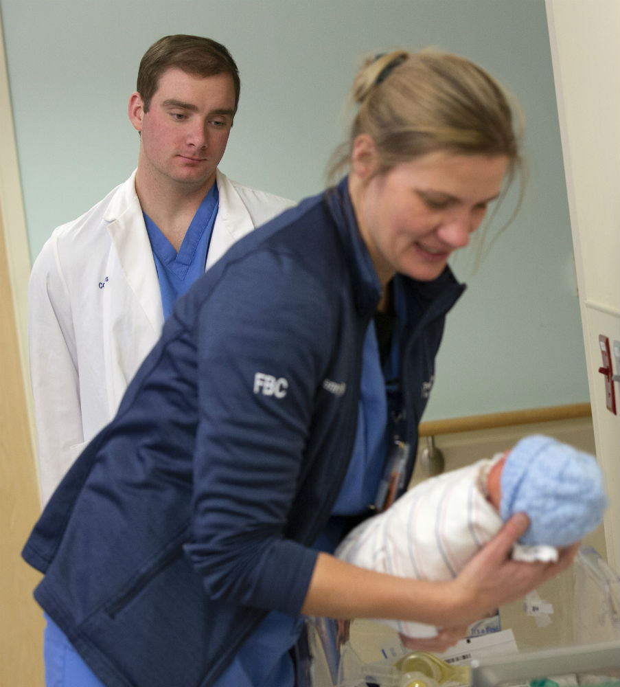 Colin Hayes watches as Jenny Meincke, a registered nurse at Penn State Health St. Joseph Medical Center, leans over while holding a newborn baby. Hayes is wearing a white coat and scrubs. Meincke is wearing a jacket and scrubs. The baby is wrapped in a blanket and has a blue knit cap on his head.