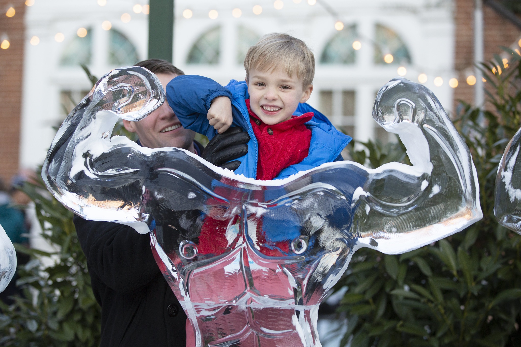 A man holds his five-year-old son up in front of an ice sculpture of a strongman making muscles at Lititz Fire and Ice on Feb. 14. The boy is smiling and wearing a sweater and coat. The man is smiling and wearing a coat and winter gloves.