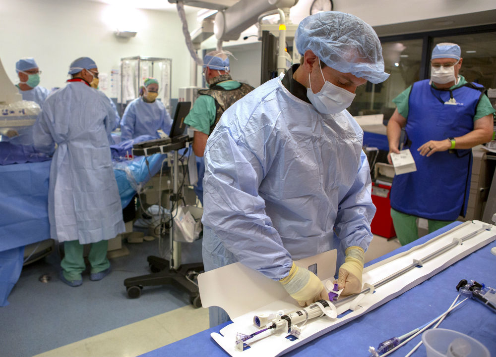 A team of six people from Penn State Heart and Vascular Institute surgeon are in an operating room and preparing for a TAVR procedure. Each are wearing surgical masks, caps and gowns. The man in the foreground is touching a heart catheter device that is lying on a table. Behind him four providers stand around the patient.