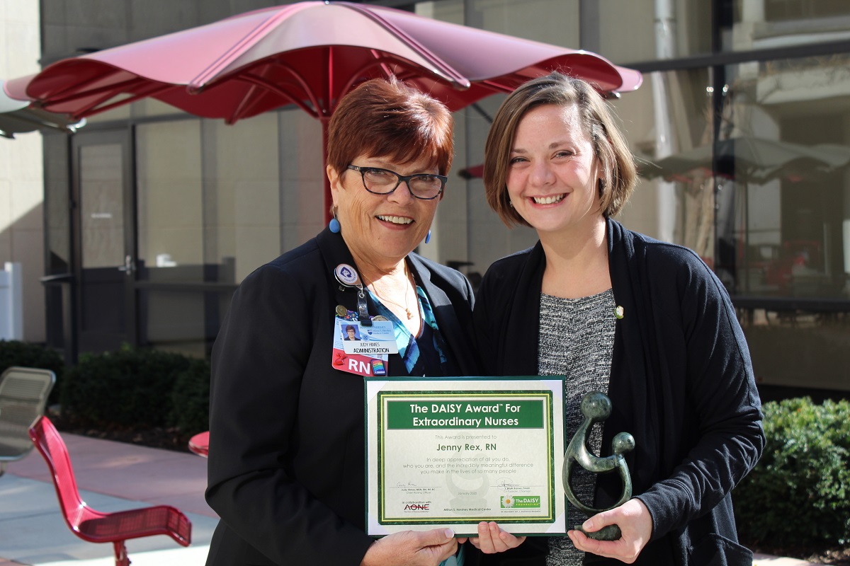 Judy Himes and Jenny Rex stand side by side and hold a plaque. Behind them is an umbrella. Rex holds a sculpture.
