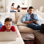 A man and young boy each use electronic devices while on a couch. A woman and young girl are in the background, slightly out of focus, sitting at a table using devices.