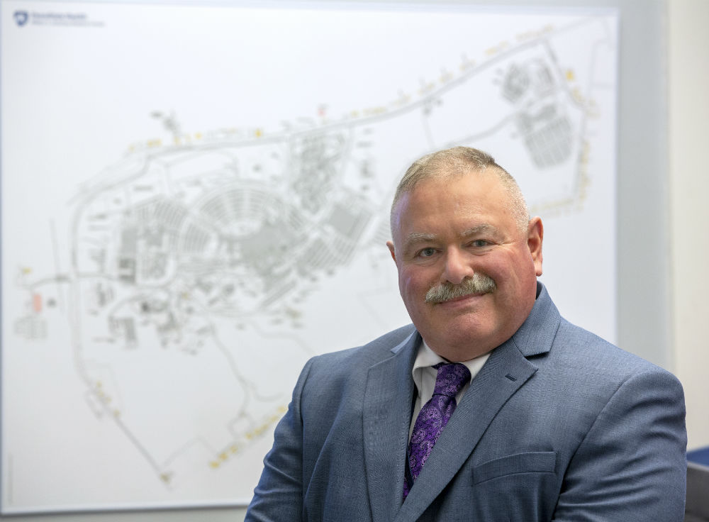 Hershey Medical Center security director Bill Losefsky sits at a desk in front of a map. He is wearing a suit, shirt and tie and has short hair and a moustache.