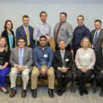 Seventeen people who are the 2019 Teacher of the Year Award recipients at the College of Medicine smile in a group photo. The back row of people are standing, and the front row are seated in chairs. A door is visible on the left.