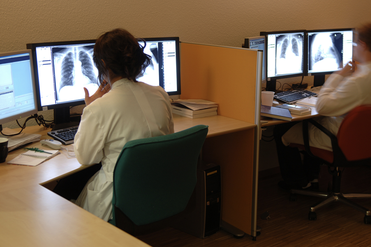 Two people in white coats are seated at computer terminals, looking at x-rays.