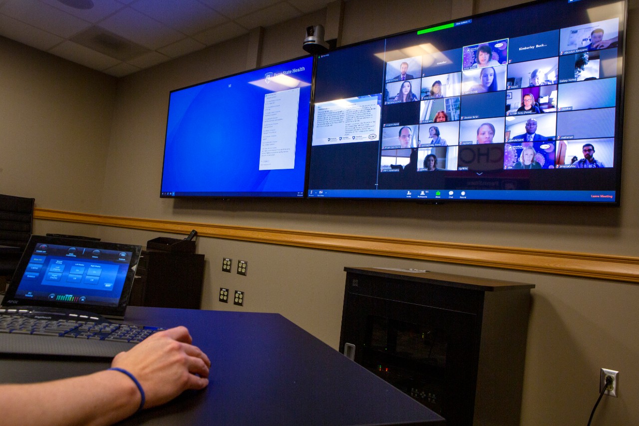 The image shows a person's hand on a desk in a room with a screen with various windows showing faces participating in an online conversation.