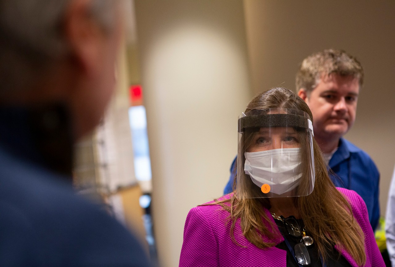 Deborah Berini stands wearing a clear shield over her face. She also wears a surgical mask. Behind her, a man, out of focus, looks on.