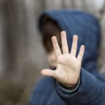 A child wearing a sweatshirt with a hood is seen out of focus and puts his hand up toward the camera.