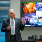Dr. David Acosta gestures while standing in front of a TV monitor loaded with out-of-focus images. Acosta wears a suit and a microphone is clipped to his tie.