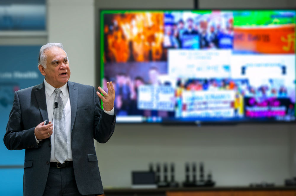 Dr. David Acosta gestures while standing in front of a TV monitor loaded with out-of-focus images. Acosta wears a suit and a microphone is clipped to his tie.