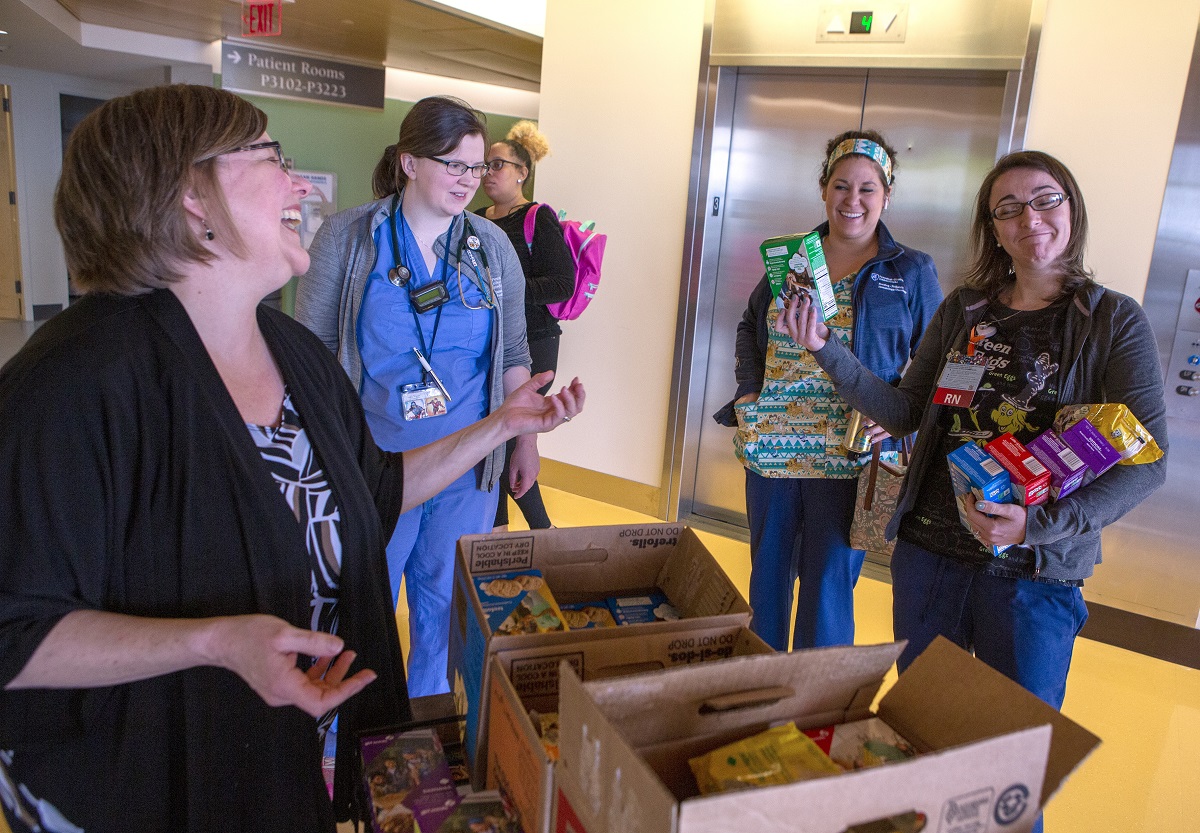 Jill Rebuck laughs at a smiling woman in scrubs holding a stack of Girl Scout cookie boxes. Other women in scrubs look on.