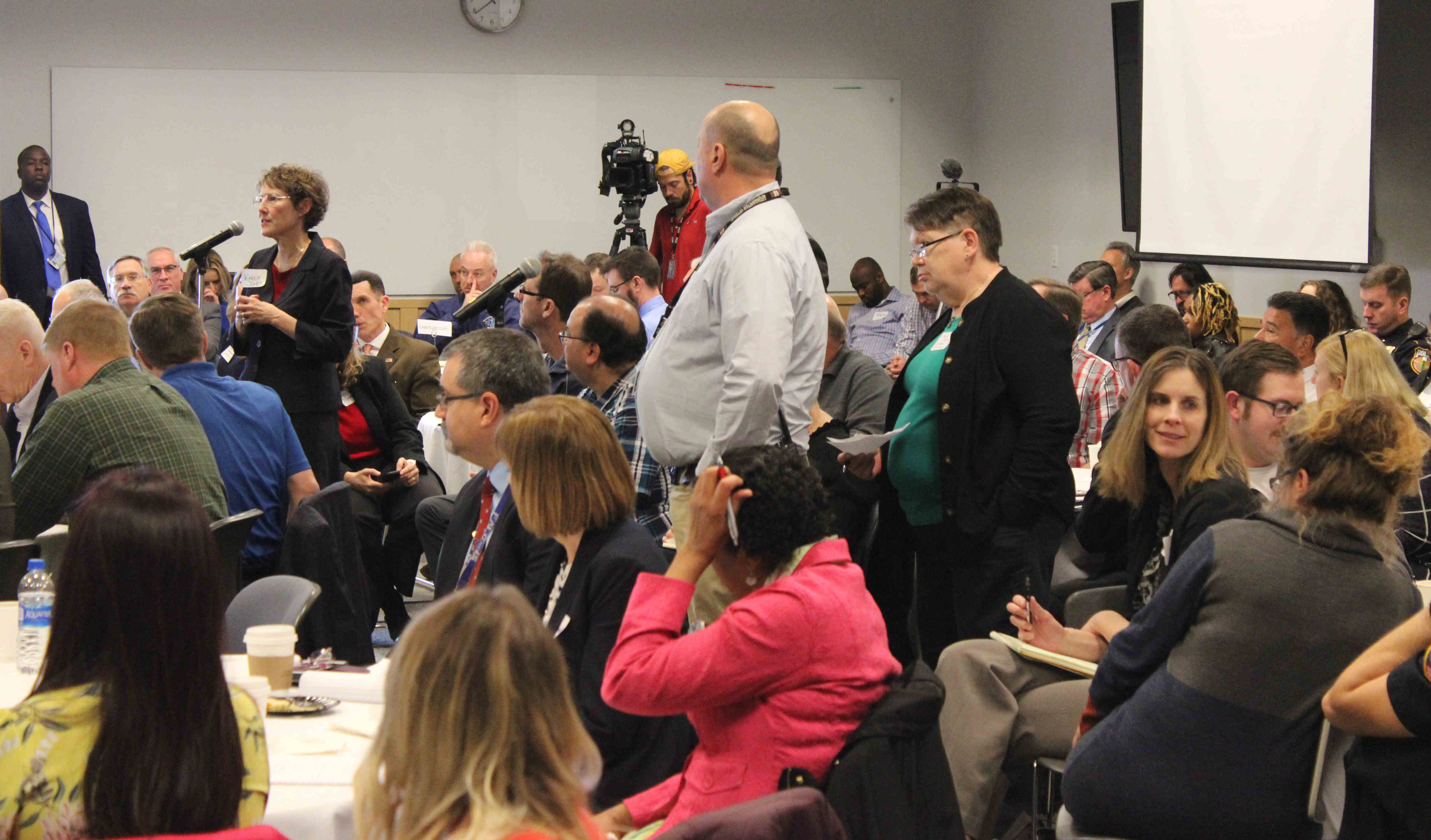 Several people are seated, all facing the same direction. A few people are standing at microphones placed in the middle of the crowd.