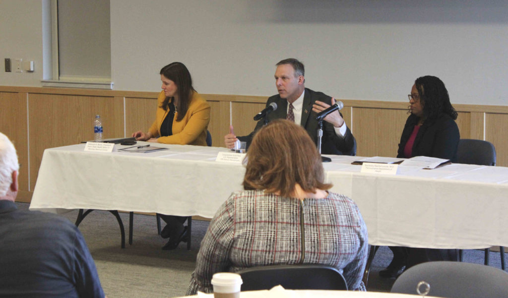 Three people – two women and a man – are seated at a table, facing an audience. The man seated in the middle, is speaking and gesturing with his hand.