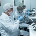 Three people sit in a row assembling personal protective face shields at Universal Protective Packaging in Mechanicsburg. They are wearing smocks, hairnets and beard nets and are holding long strips of paper.