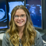 Kassie Heeman, a sonographer at Hershey Medical Center, smiles as she sits in front of desks with four monitors. The monitors have ultrasound images on them. Kassie has long hair and is wearing glasses and a jacket with a Penn State Health logo on it.