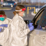 Two nurses wearing personal protective gear administer a car side test for COVID-19