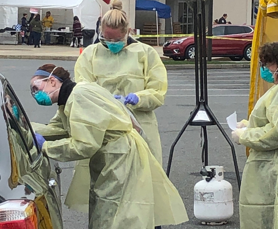 Nurses wearing personal protective equipment help a patient next to a vehicle at a drive-through testing site