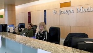 Khaliah Morris, left, and Lori Munoz sit at the front desk of St. Joseph Medical Center. They are smiling and wearing sweatshirts. The St. Joseph logo is behind them on the wall.