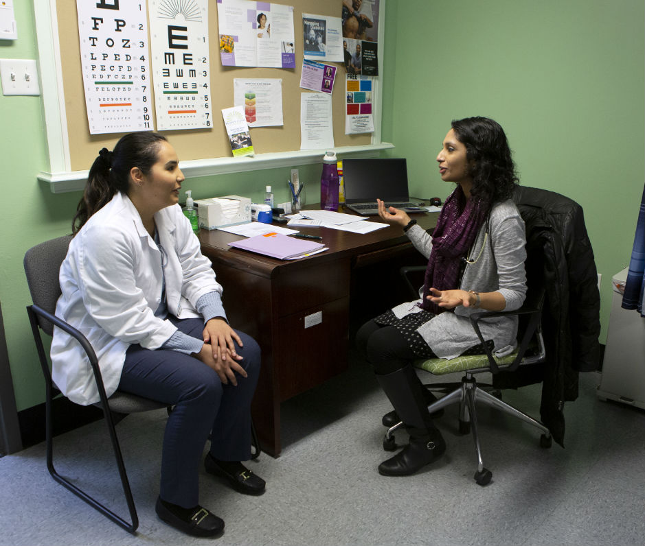 Dr. Leesha Helm, seated at a desk, gestures toward Penn State College of Medicine third-year student Maria Escudero during a shadowing visit at Beacon Clinic. Escudero, who sits in a chair by the desk, wears her white coat over pants and has a long ponytail. Helm, who has long wavy hair, wears a long sweater and scarf over leggings. A bulletin board with an eye chart and other papers on it is on the wall behind the desk, which has papers and a laptop on it.