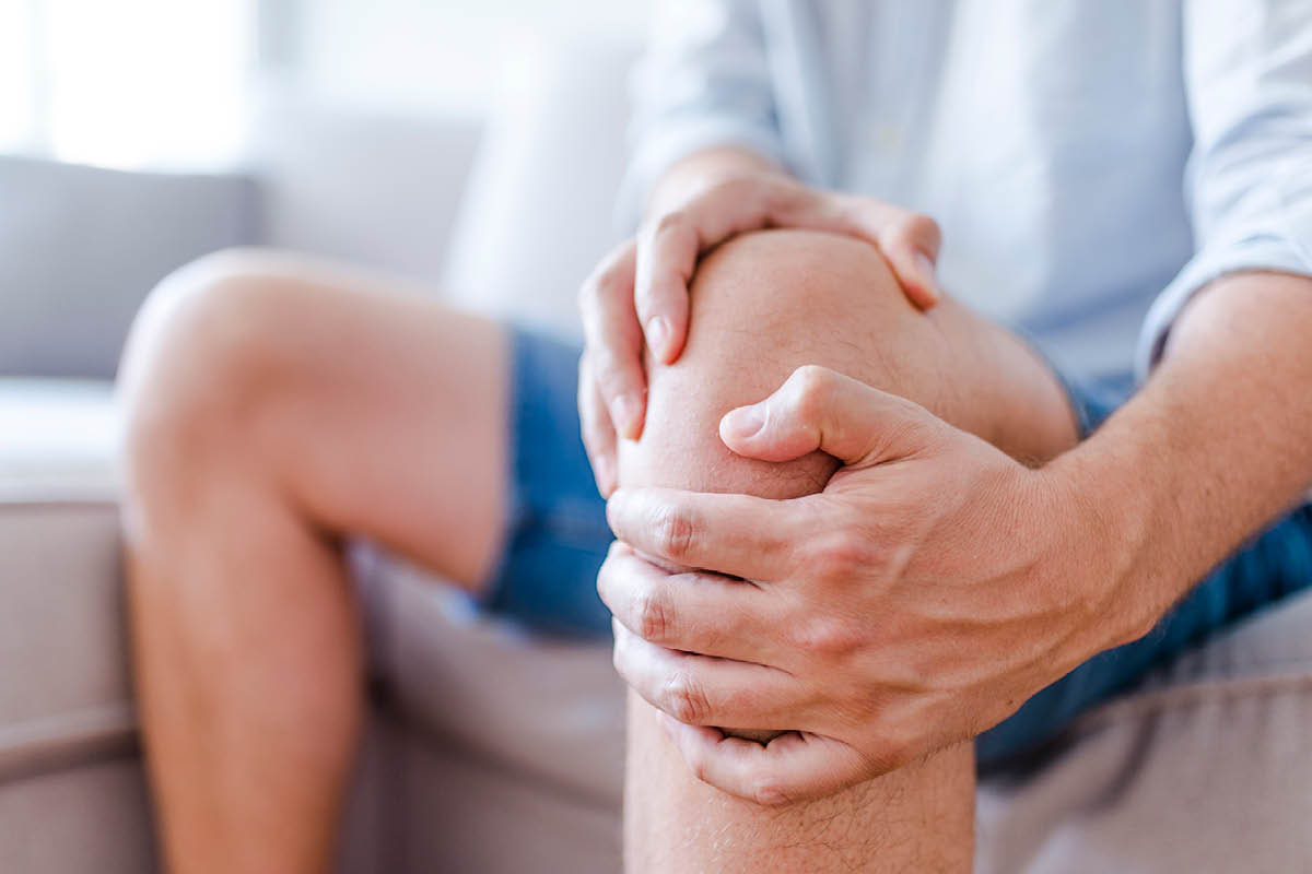 Close-up shot of a man, seated on a couch, holding his left knee with both hands.