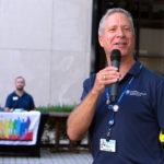 A man speaks into a microphone at an outdoor event. Two people are seen in the background at a table of LGBTQ+ information.