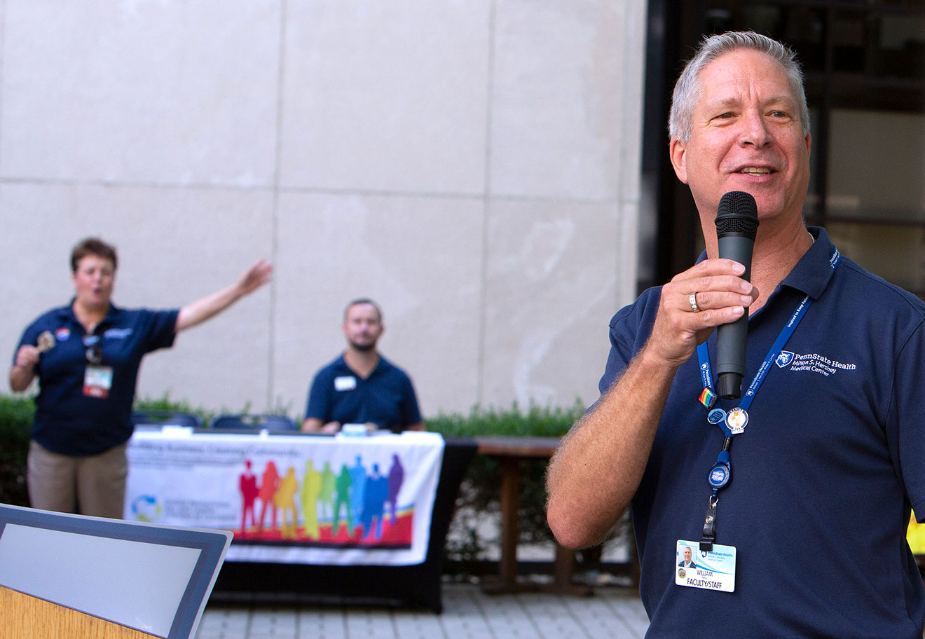 A man speaks into a microphone at an outdoor event. Two people are seen in the background at a table of LGBTQ+ information.