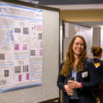A woman stands near an academic poster, talking and smiling to another person.