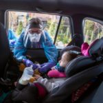 Penny Porter a nurse practitioner with Penn State Health Medical Group ― All About Children, leans into a car and touches the fingers of 2-year-old Zyann Perez-Cruz. Porter is wearing a gown and face shield. The child is in a car seat and has a bottle in the cup holder next to her.