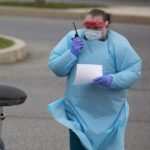 A nurse wearing a gown, gloves and a face shield holds a walkie-talkie as she stands next to a woman in a car. The woman is wearing a face mask.