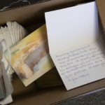 Stacks of cards sit in a cardboard box. The front of the card shows a watercolor painting of Hershey Medical Center. One card is propped open to show handwriting inside.