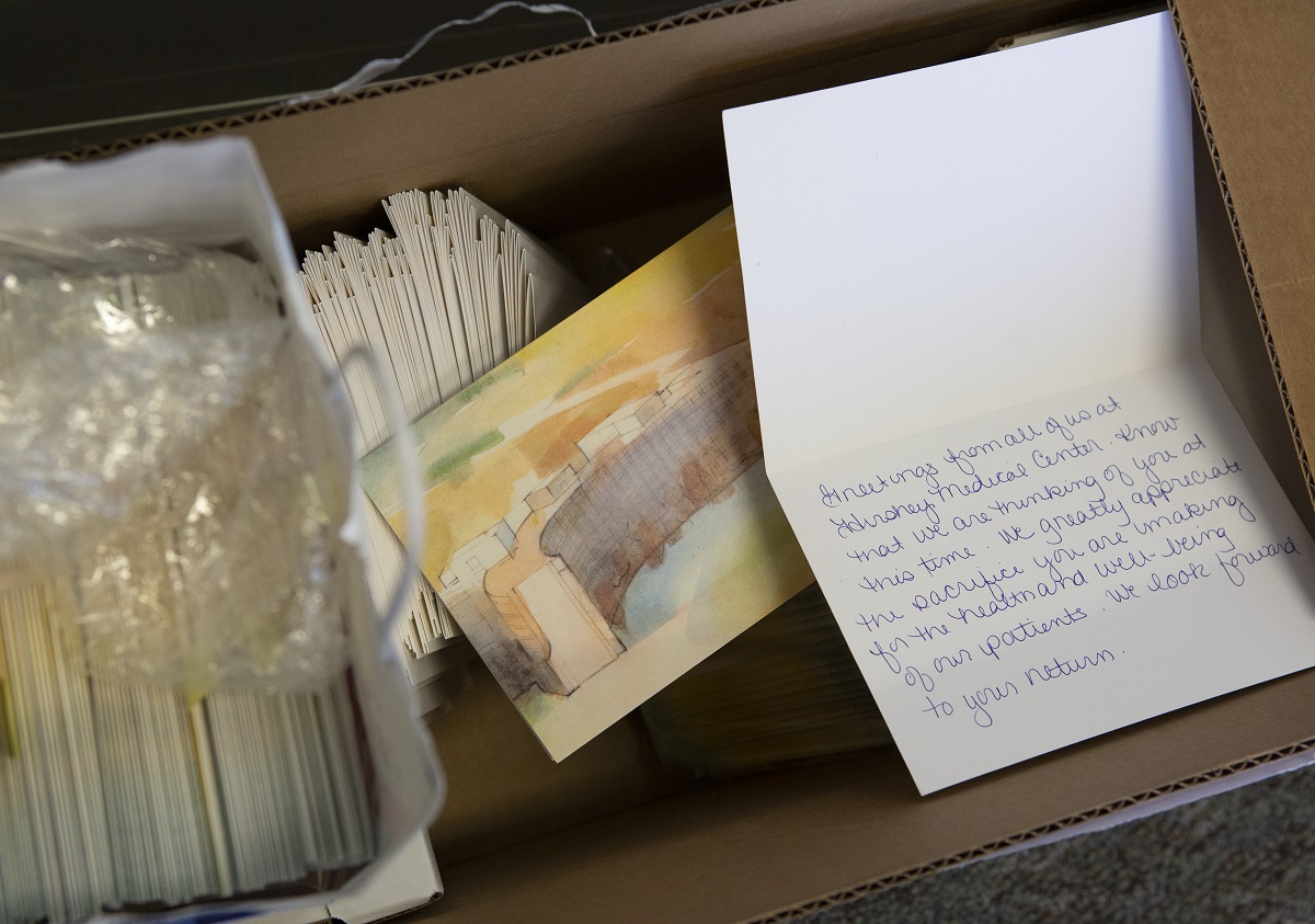 Stacks of cards sit in a cardboard box. The front of the card shows a watercolor painting of Hershey Medical Center. One card is propped open to show handwriting inside.