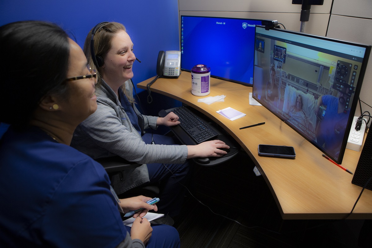 Jennifer Gish, nurse manager, and registered nurse Marilou Magnaye sit at a curved desk and smile at a computer monitor that shows stroke patient Kimberly Laughman in her hospital room in bed, with nurses and equipment around her.