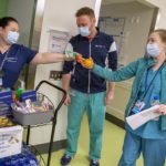 Nurse manager Stephanie Reed stands behind a two-shelf cart filled with fruit, packaged snacks and bottled water, and hands a candy bar and orange to Dr. Holly Whitt, who reaches her hand out to take the chocolate. Dr. Robert Kavanaugh, dressed in scrubs, stands between them with his arms at his sides. They are all wearing face masks.