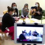 Three Penn State Health Children's Hospital staff members sit around a child-sized table, coloring with crayons and paper while music therapist Devon Springer strums a guitar. They are all wearing masks. In the foreground is a small screen showing the scene as it is recorded.