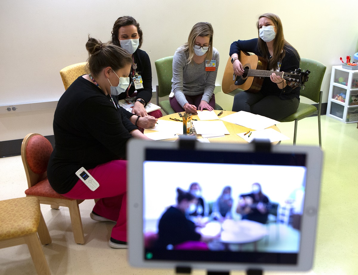 Three Penn State Health Children's Hospital staff members sit around a child-sized table, coloring with crayons and paper while music therapist Devon Springer strums a guitar. They are all wearing masks. In the foreground is a small screen showing the scene as it is recorded.