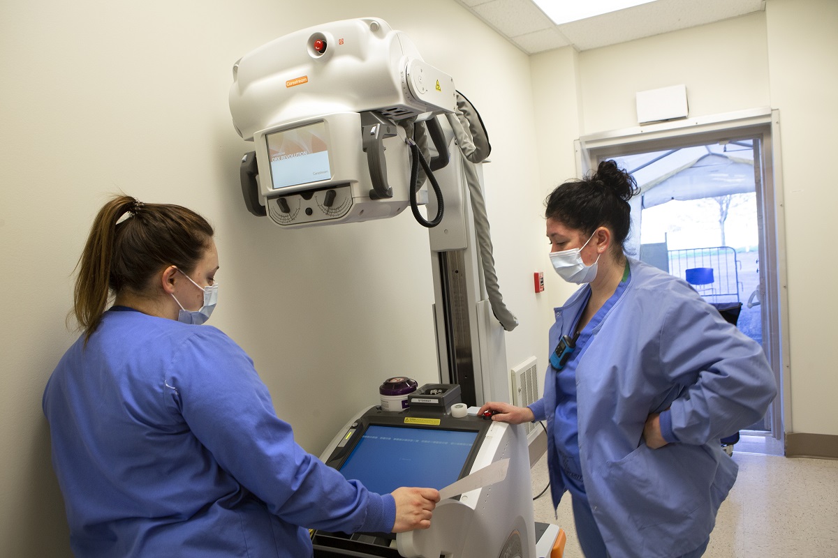 Two radiologic technologists at Hershey Medical Center set up a portable X-ray machine. They are standing by the machine wearing face masks and scrubs. The machine has a large camera on a stand.