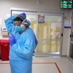 A health care worker at Hershey Medical Center wearing personal protective equipment adjusts her face shield. Behind her are two glass doors and a medical waste container.