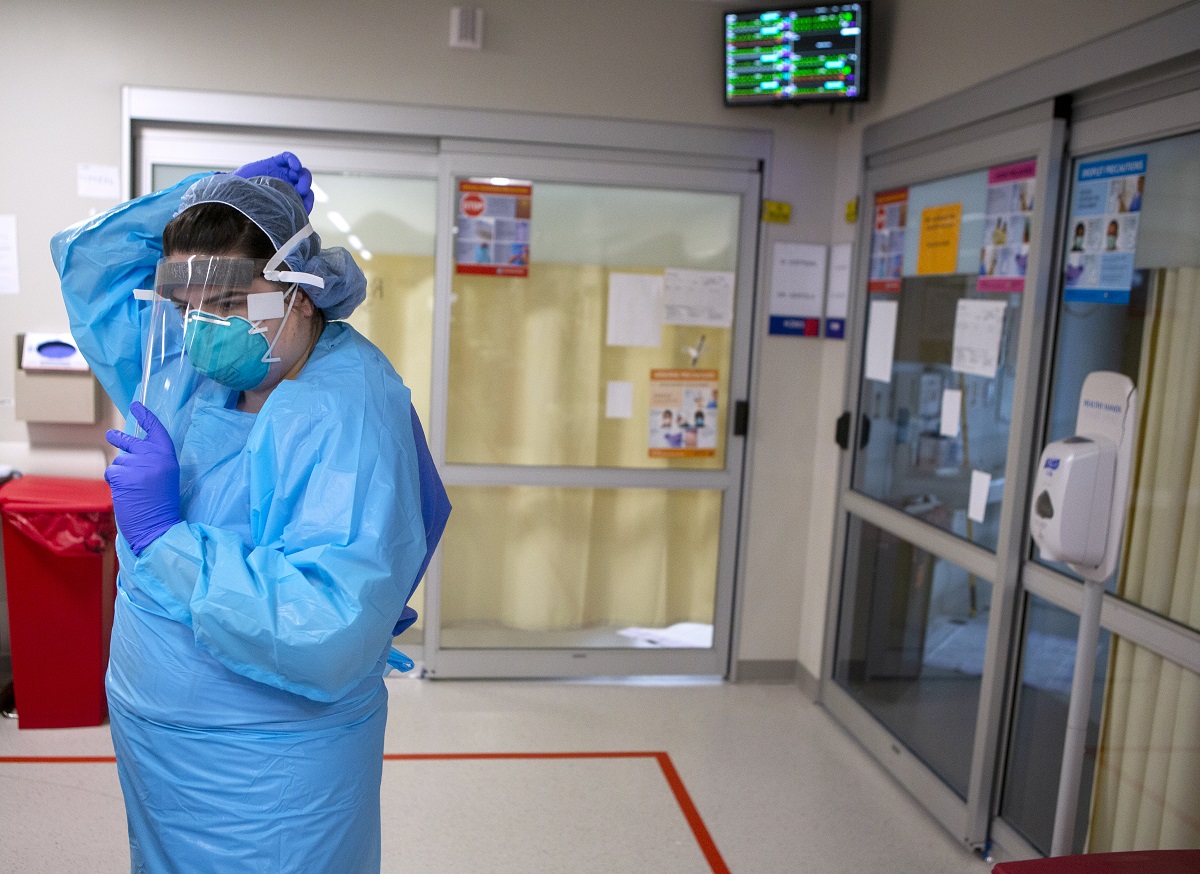 A health care worker at Hershey Medical Center wearing personal protective equipment adjusts her face shield. Behind her are two glass doors and a medical waste container.