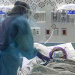 Dr. Jennifer Cooper, wearing a gown and face shield, leans over a female patient who is lying in bed wearing a surgical cap and face mask. Behind them are a monitor and medical equipment attached to the wall.