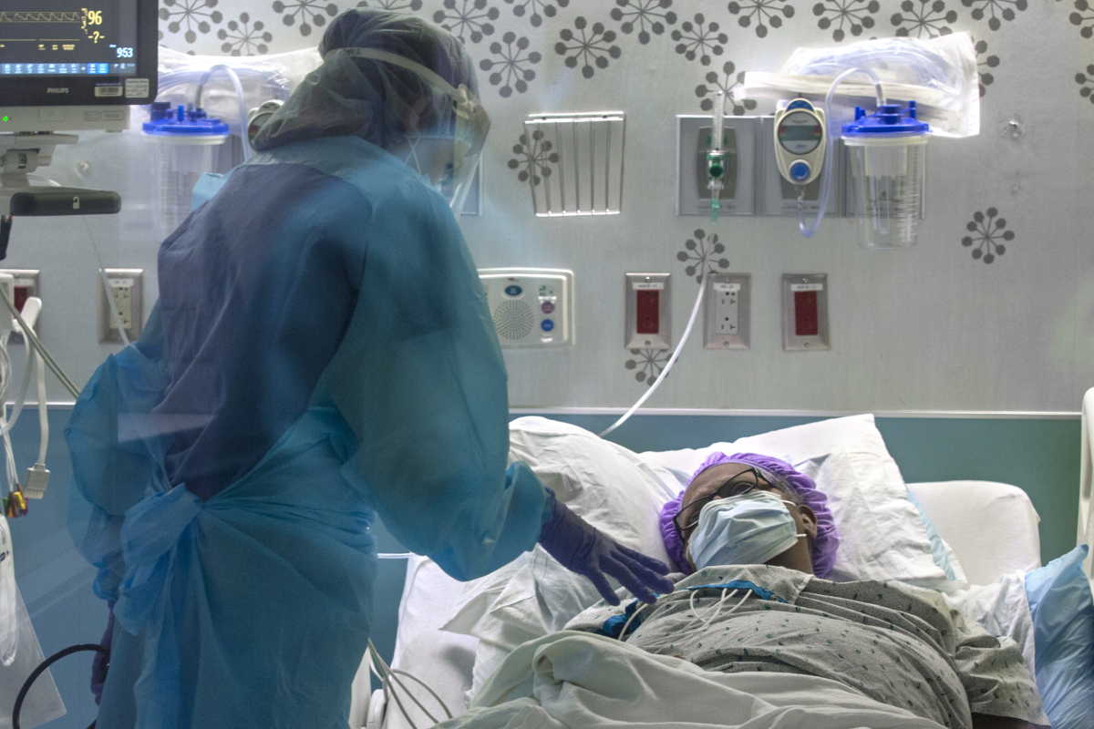 Dr. Jennifer Cooper, wearing a gown and face shield, leans over a female patient who is lying in bed wearing a surgical cap and face mask. Behind them are a monitor and medical equipment attached to the wall.