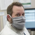 A man with a beard wears a cloth face mask and a jacket and looks at the camera. Behind him are two computer monitors and a bulletin board.