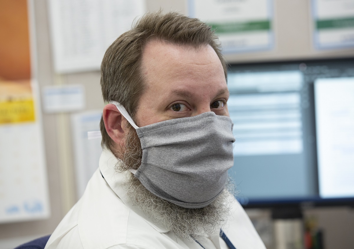 A man with a beard wears a cloth face mask and a jacket and looks at the camera. Behind him are two computer monitors and a bulletin board.