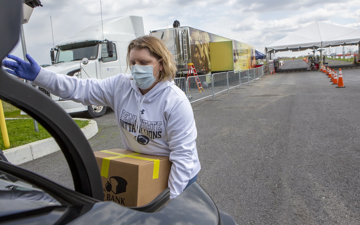 Registered nurse Stephanie McGaw, who is wearing a sweatshirt, gloves and a mask, lifts a cardboard box of food into the trunk of a car. A tractor-trailer truck and traffic cones marking drive-through lanes are in the parking lot behind her.