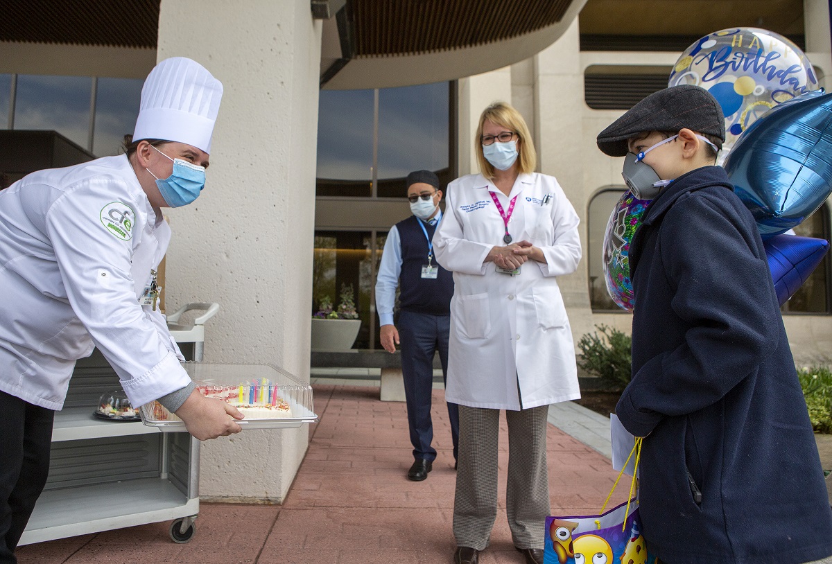 A chef wearing a face mask holds a cake out to a boy who is wearing an N95 mask, a coat and hat. Behind him are balloons, a woman in a white coat and a man.